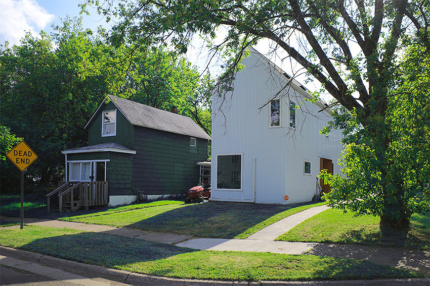 Two contrasting houses on a sunny day: a rustic green one with a porch and a modern white one with large windows, both narrow and with pitched roofs.