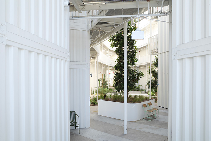 A courtyard with lush plantings surrounded by the angled white walls of container modules and open-air walkways leading to apartment doors.