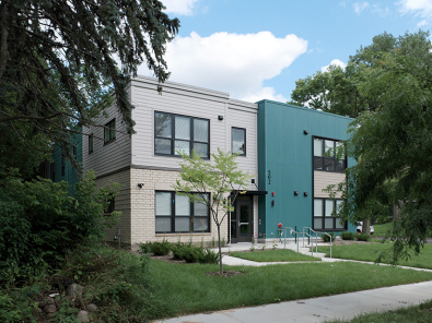 A modern two-story apartment building with teal and beige exterior siding, large windows with black trim, and a flat roof.