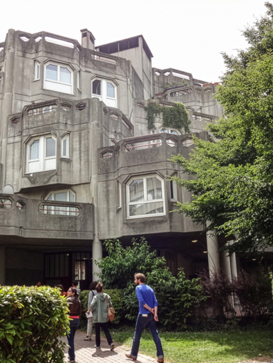 People walking into a multilevel building with geometric designs, surrounded by greenery.