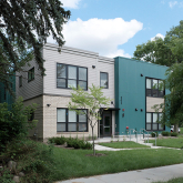 A modern two-story apartment building with teal and beige exterior siding, large windows with black trim, and a flat roof.