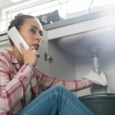 Woman on phone with hand on leaking sink pipe.