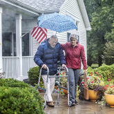 Older couple walking in front of a house, one with a walker