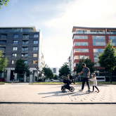 Father, child, and baby in a stroller walk past two multifamily buildings