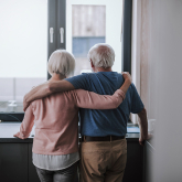 Older adult couple looking out of apartment window.
