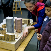 Two women and a child point discuss a model of the proposed redevelopment. New buildings show windows and balconies. Existing buildings are shown in plexiglass.