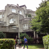 People walking into a multilevel building with geometric designs, surrounded by greenery.