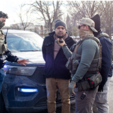 Masked immigration agents surround an arrested man in handcuffs.