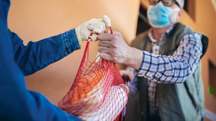 Gloved person hands bag of groceries to masked senior man.
