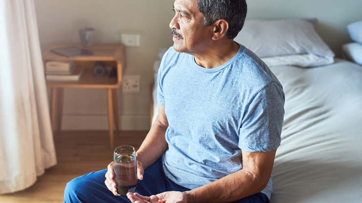 Older man sitting in bedroom with medication and glass of water.
