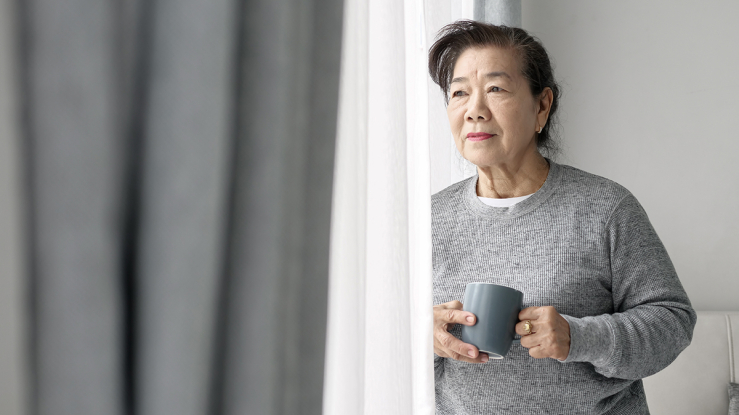Older woman looking out window while holding mug.
