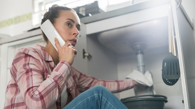 Woman on phone with hand on leaking sink pipe.