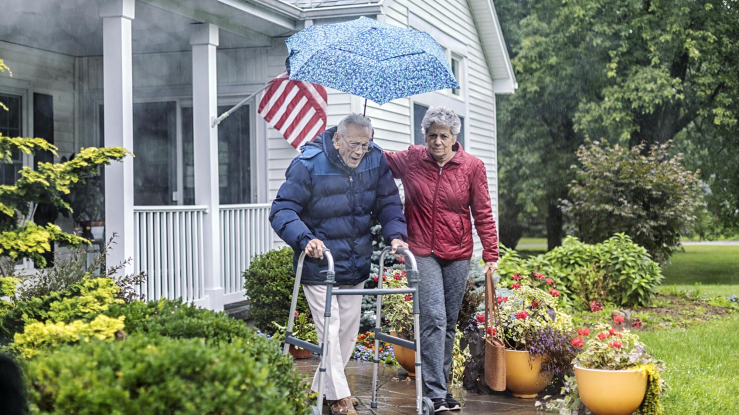 Older couple walking in front of a house, one with a walker