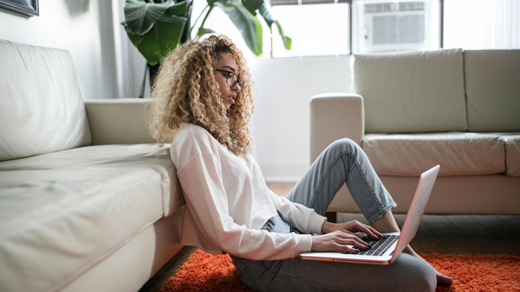 Young woman sitting on floor by couch with laptop.