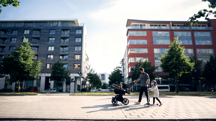 Father, child, and baby in a stroller walk past two multifamily buildings