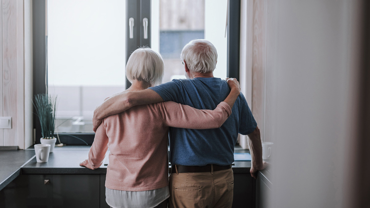 Older adult couple looking out of apartment window.