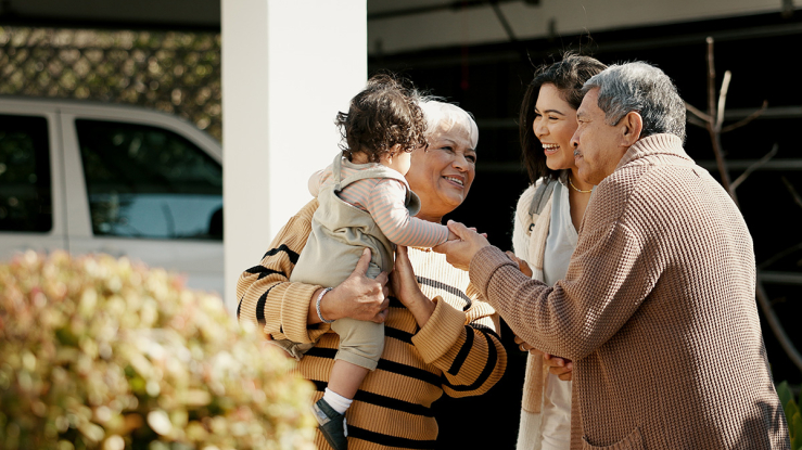 Older Hispanic couple greeting daughter and grandchild outside their home.