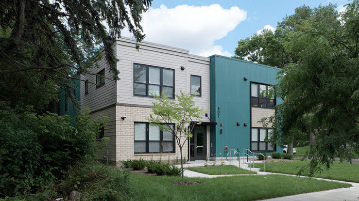 A modern two-story apartment building with teal and beige exterior siding, large windows with black trim, and a flat roof, surrounded by lush trees and grass.