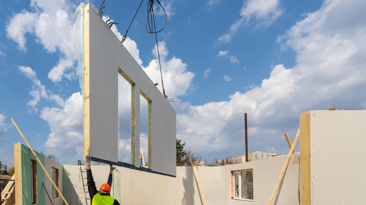 Crane placing prebuilt wall into house under construction.