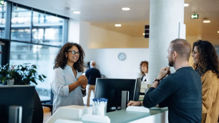 Woman at reception desk welcoming two visitors in an office lobby.