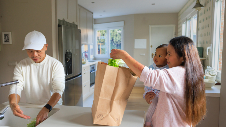 Woman holding a child while unpacking groceries at home.