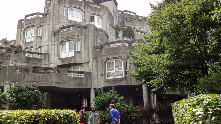 People walking into a multilevel building with geometric designs, surrounded by greenery.