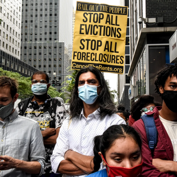 Crowd of protestors in urban area with "stop all evictions, stop all foreclosures" sign.