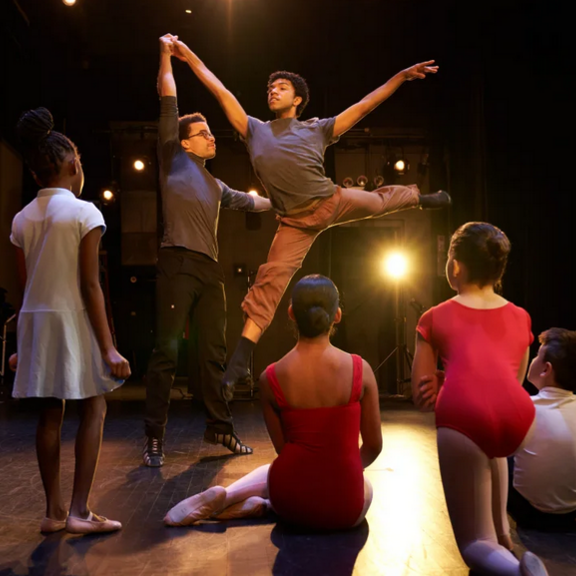 Dancer leaping with guidance from another dancer, with students watching.