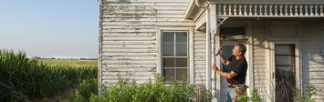 View of a man hammering a nail on the porch of an old house.