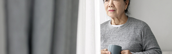 Older woman looking out window while holding mug.