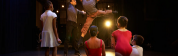 Dancer leaping with guidance from another dancer, with students watching.